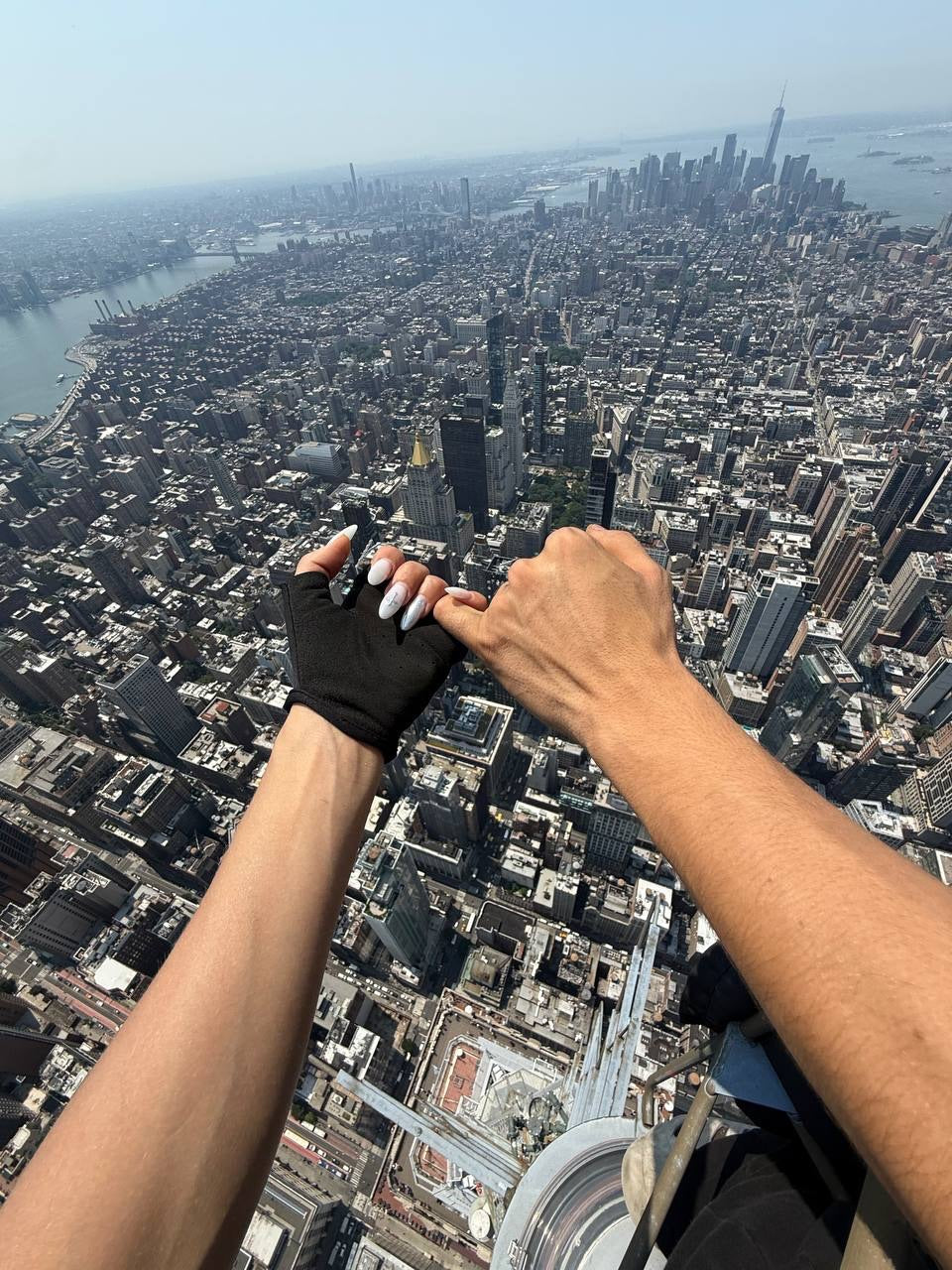 Two people high above a dense city skyline, with one person wearing a black fingerless glove and the other holding their hand. The background shows a sprawling urban landscape with skyscrapers, water, and bridges under a hazy sky.