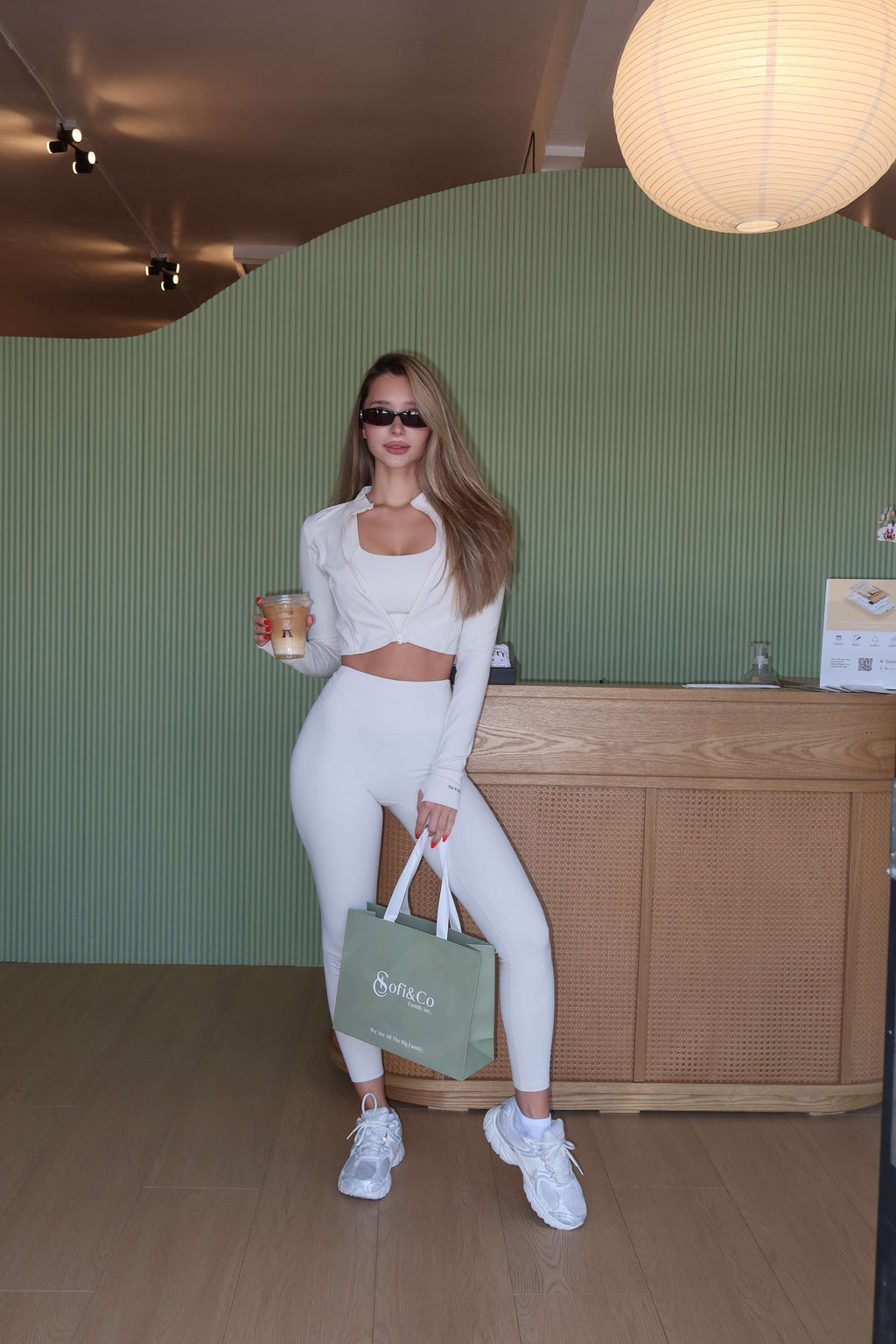 A woman posing in a cafe or boutique setting, wearing a matching white long-sleeve crop top and high-waisted leggings set, white sneakers, and holding a green tote bag with the brand name 'SimpCo'.
