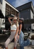 Two women posing outdoors in front of a modern building labeled 'west elm' and 'MANHATTAN AVE'. The woman on the left wears a white crop top and beige leggings, while the woman on the right wears a black top, a patterned beanie, and light-wash blue jeans.
