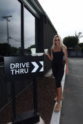 A woman in a black dress stands outdoors next to a black 'DRIVE THRU' sign, holding takeaway coffee cups near a modern building with large windows.