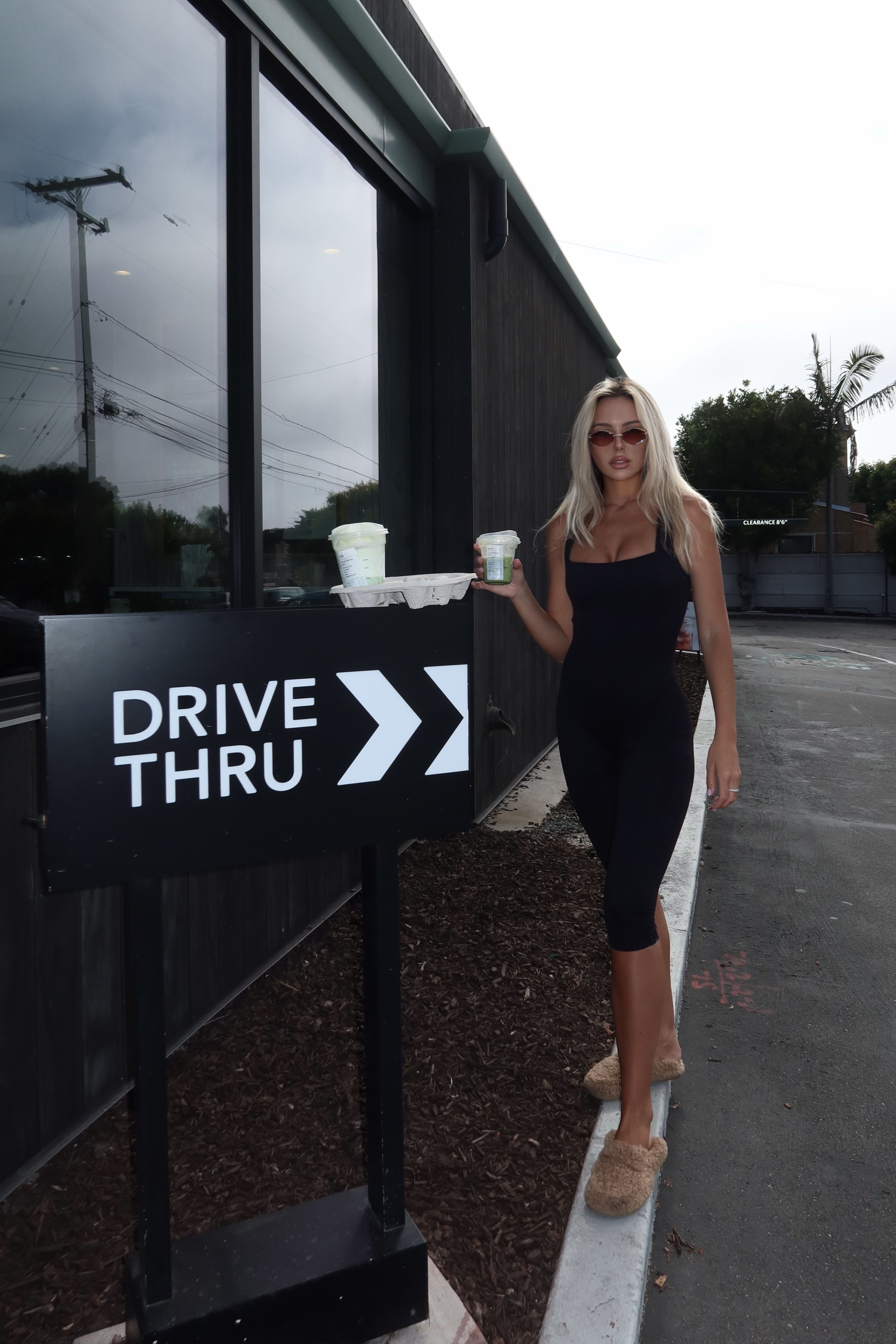 A woman in a black dress stands outdoors next to a black 'DRIVE THRU' sign, holding takeaway coffee cups near a modern building with large windows.