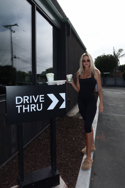 A woman in a black dress stands outdoors next to a black 'DRIVE THRU' sign, holding takeaway coffee cups near a modern building with large windows.