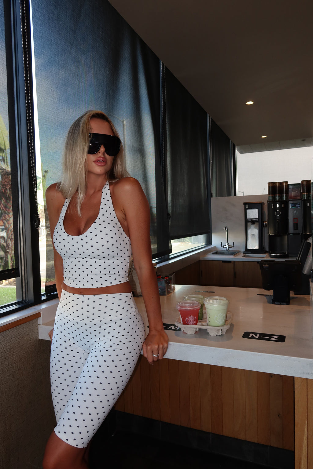A woman wearing a white two-piece halter crop top and matching knee-length skirt with black polka dots, posing in a modern cafe or restaurant setting.