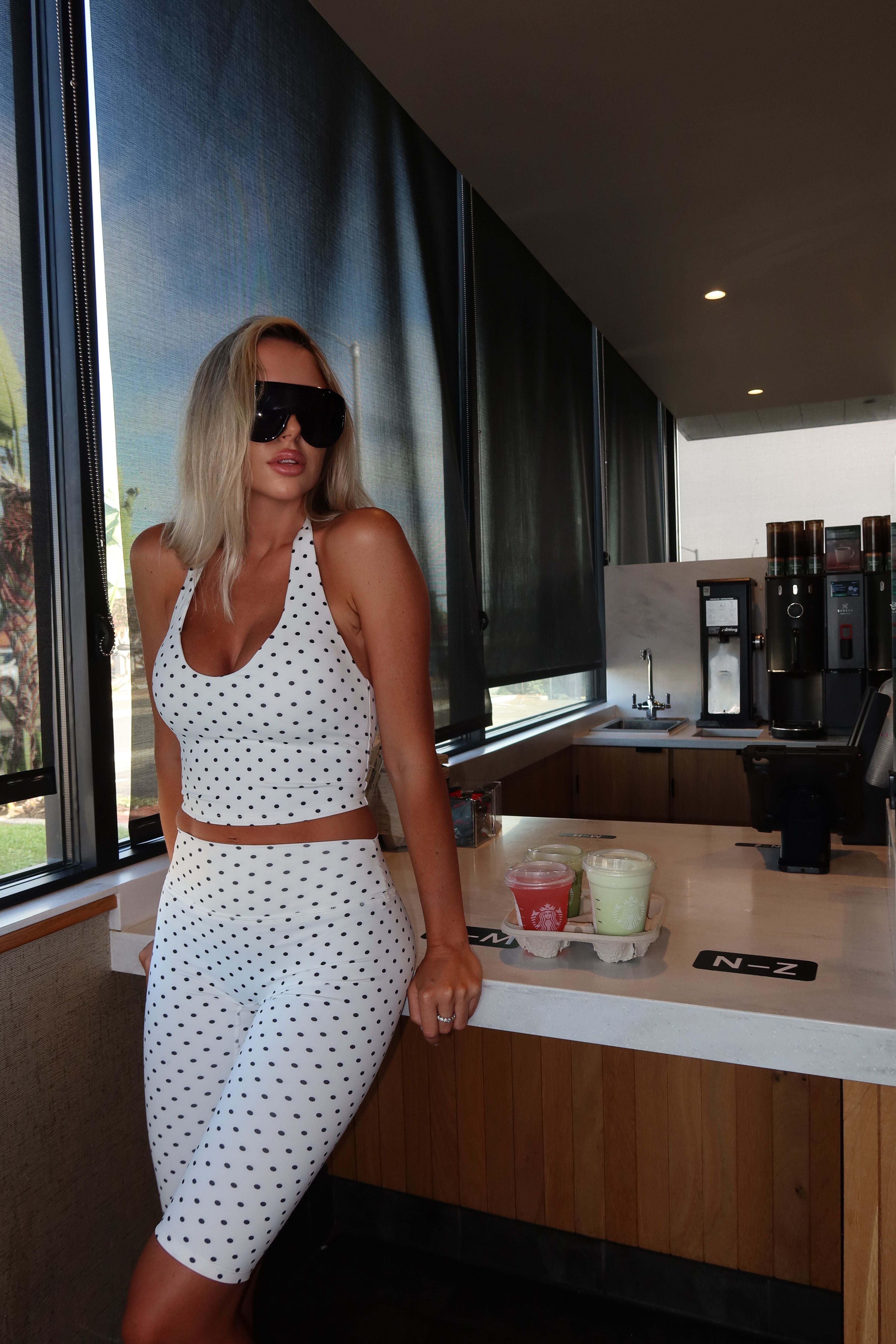 A woman wearing a white two-piece halter crop top and matching knee-length skirt with black polka dots, posing in a modern cafe or restaurant setting.