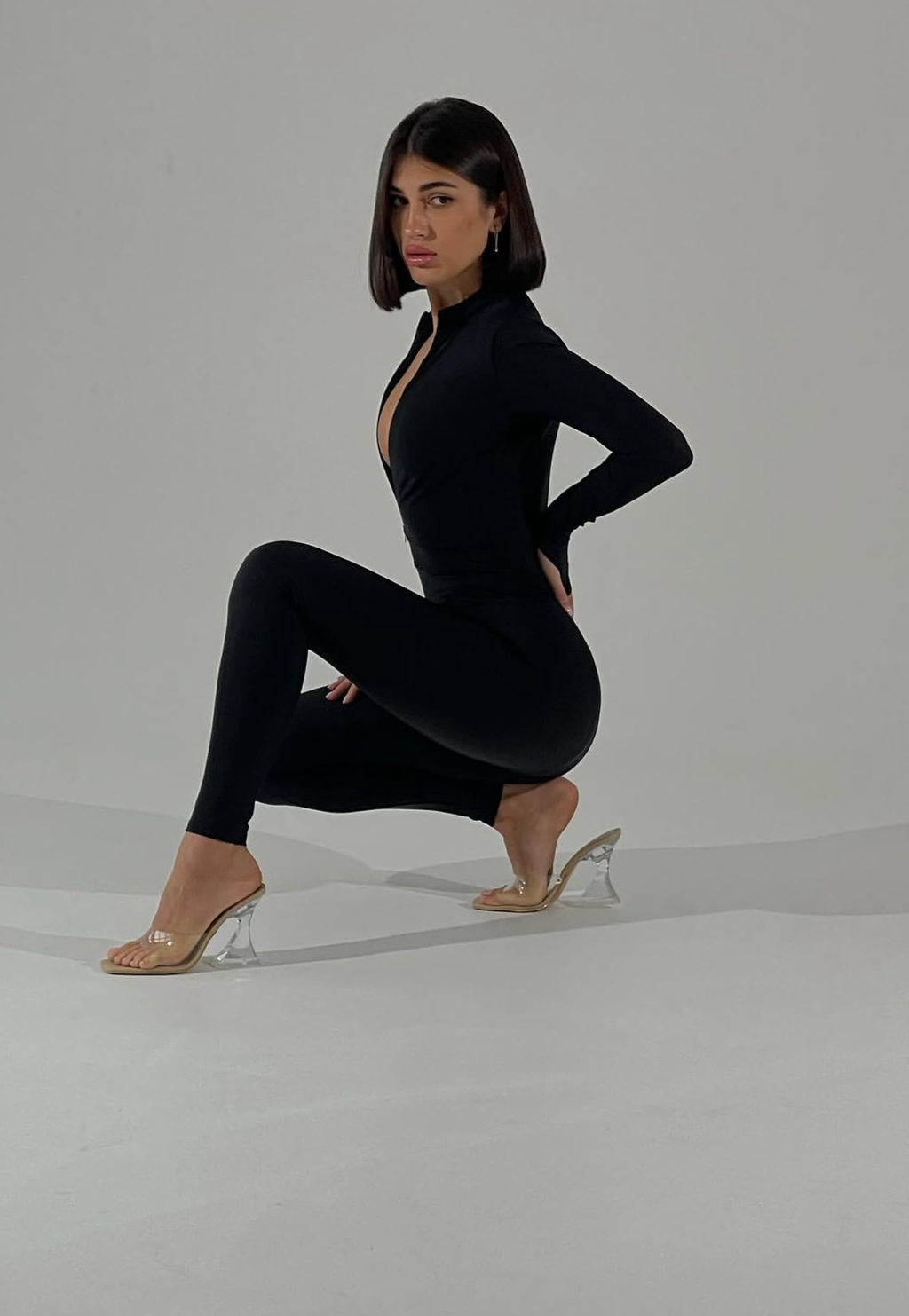 A woman posing in a black long-sleeve bodysuit with a plunging neckline and matching leggings, paired with clear high-heeled sandals, set against a plain light gray studio background.