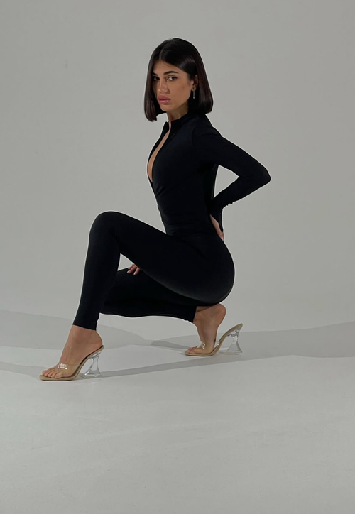 A woman posing in a black long-sleeve bodysuit with a plunging neckline and matching leggings, paired with clear high-heeled sandals, set against a plain light gray studio background.