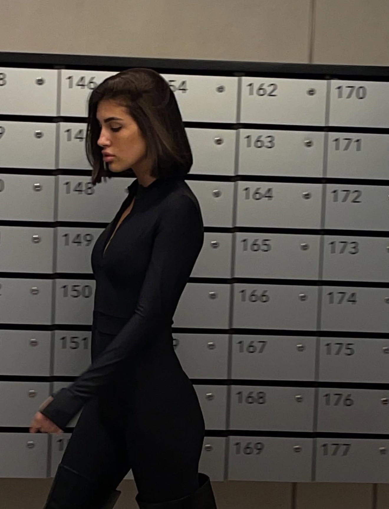 A woman with shoulder-length dark brown hair walks in profile wearing a fitted black long-sleeve top and black pants, standing in front of a row of gray metal lockers numbered 146 through 177.