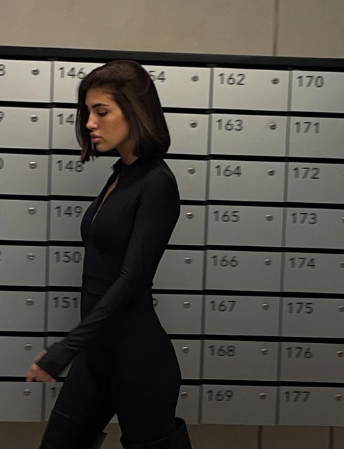 A woman with shoulder-length dark brown hair walks in profile wearing a fitted black long-sleeve top and black pants, standing in front of a row of gray metal lockers numbered 146 through 177.